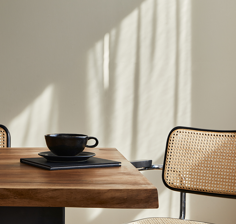 Warm dining room interior with wooden family table, rattan design chairs, cup of coffee with natural window light casting shadows on the wall behind